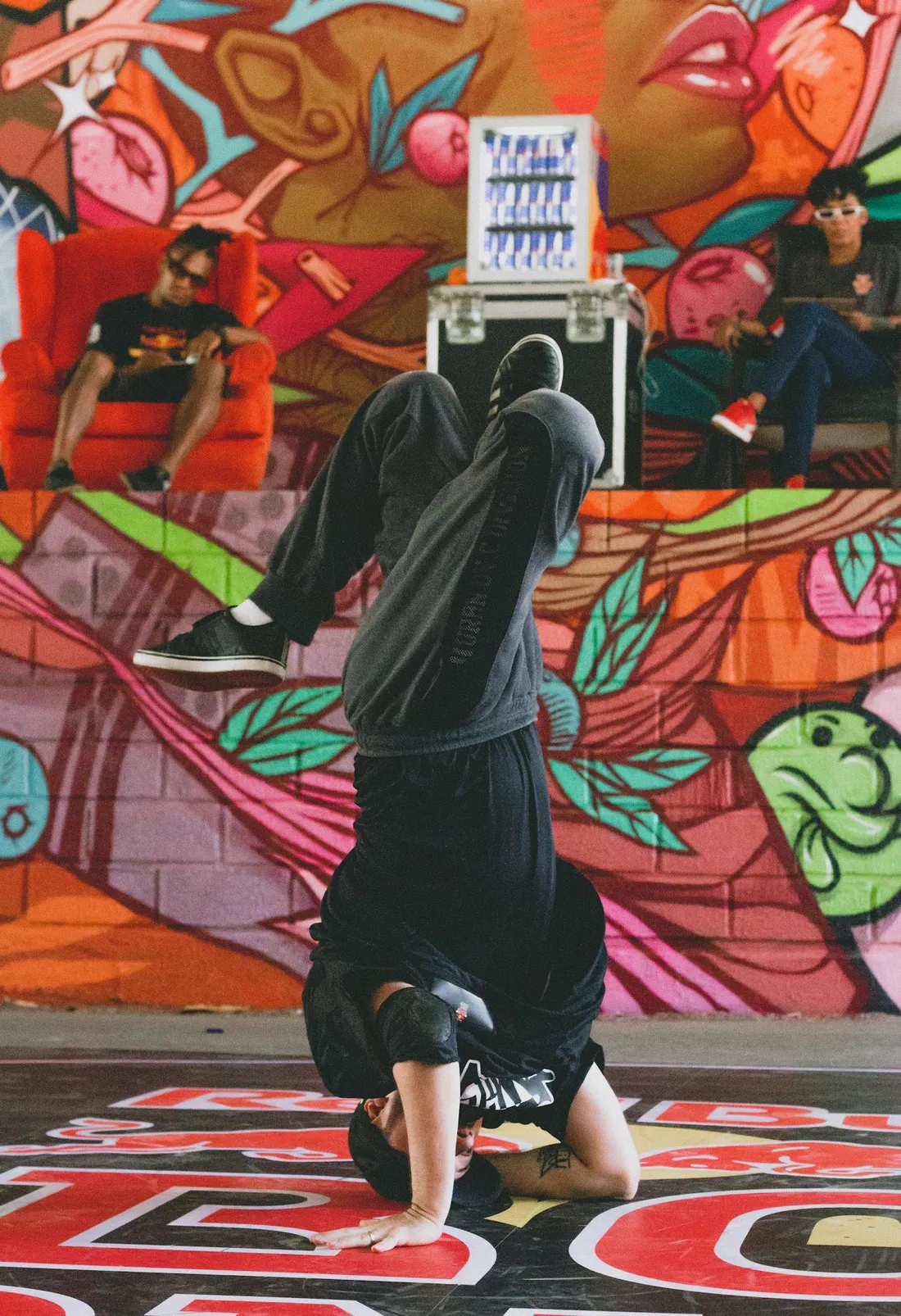 A breakdancer executes a headstand in front of vivid graffiti art while spectators look on.